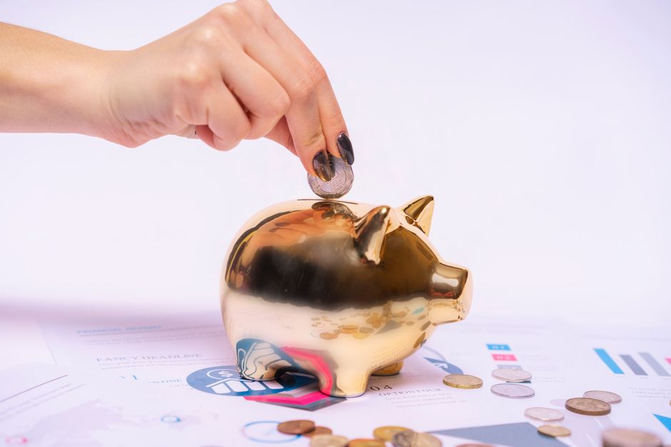 A close-up of a hand placing a coin into a shiny, gold-colored piggy bank, positioned on a table with financial documents, charts, and additional scattered coins. The background is plain and light-col